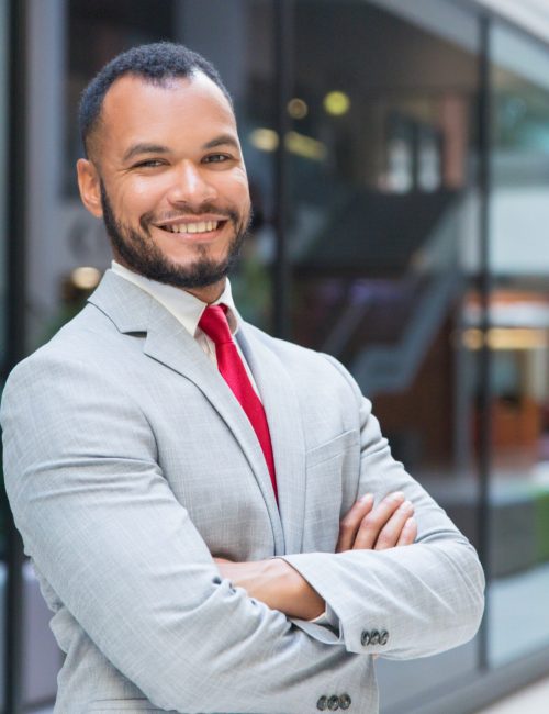 Cheerful businessman smiling at camera. Portrait of confident African American businessman in suit standing with crossed arms outside office building and looking at camera. Business concept